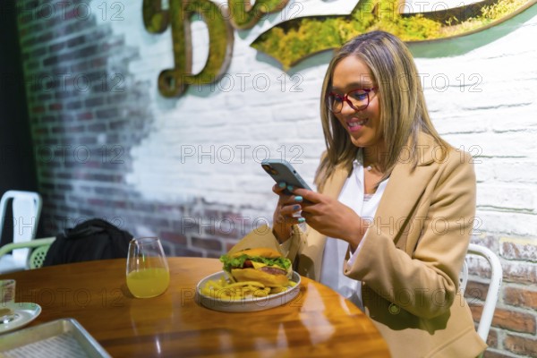 Beautiful latin woman wearing business casual clothes enjoying tasty burger and texting on the phone