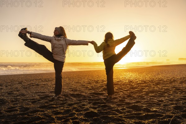 Two women are doing yoga on a beach. The sun is setting in the background, creating a warm and peaceful atmosphere