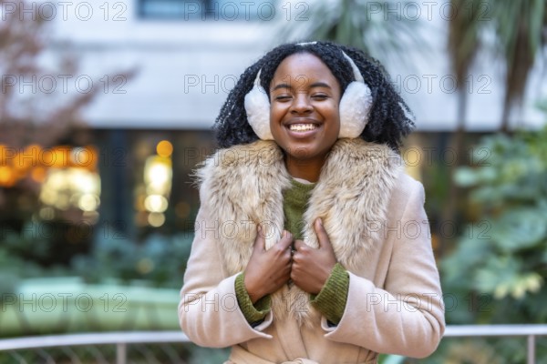 Happy young woman wearing cozy earmuffs and a stylish winter coat, smiling brightly while enjoying the vibrant city atmosphere during winter