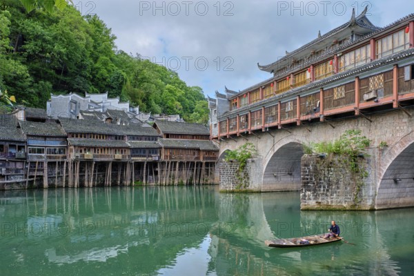 FENGHUANG, CHINA, APRIL 23, 2018: Unidentified chinese man in boat Feng Huang Ancient Town (Phoenix Ancient Town) on Tuo Jiang River with bridge and tourist boat. Hunan Province, China, Asia