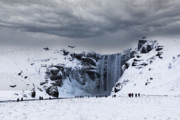 Tourists at Skogafoss waterfall, snowy and icy rock face, dark clouds above, Sudurland, Iceland, Europe