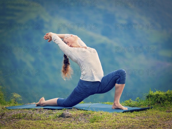 Yoga outdoors, sporty fit woman practices Hatha yoga asana Anjaneyasana, low crescent lunge pose posture outdoors in Himalayas mountains. Vintage retro effect filtered hipster style image
