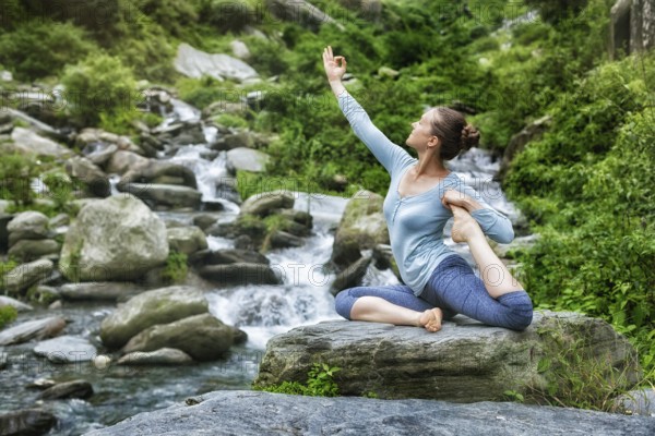 Young sporty fit woman doing yoga asana Eka pada rajakapotasana, one-legged king pigeon pose at tropical waterfall