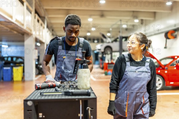 Two auto mechanics, a man and a woman, stand beside a workbench, reviewing tools and discussing car maintenance in a bustling garage