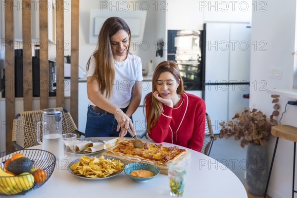 Two women are slicing and sharing a rectangular pizza, nachos with sauce and tacos at home in their modern kitchen