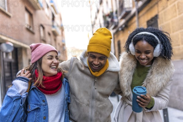 Three cheerful young multi ethnic friends are enjoying a winter day together, walking arm in arm through a city street, laughing and having a good time
