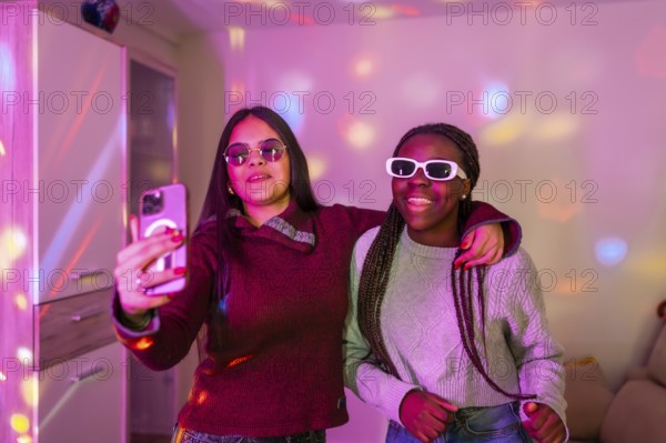 Two happy teenage girls with sunglasses taking a selfie with smartphone at a home party with disco lights