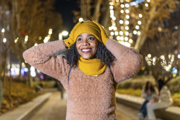 Smiling young woman wearing winter clothes adjusting her yellow wool cap under christmas lights in a city park at night