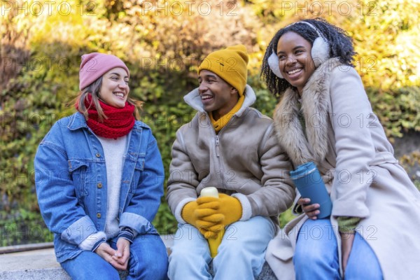 Group of young multi ethnic friends wearing winter clothes, sitting outdoors in the city and enjoying a hot beverage together on a sunny winter day