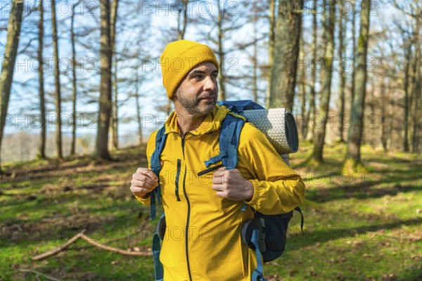 Male hiker in a bright yellow jacket and blue backpack walking through a lush forest, soaking up the sun on a beautiful day
