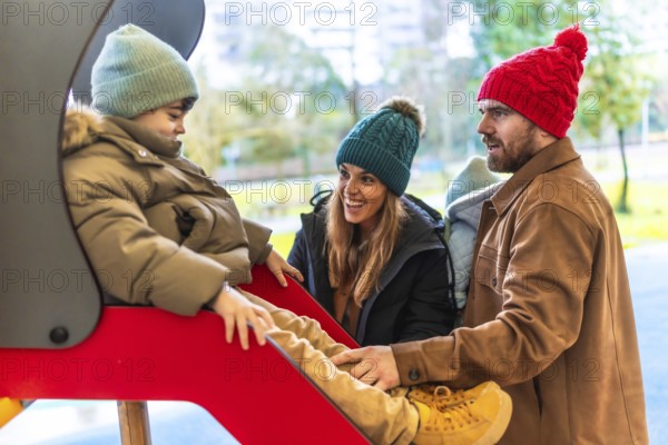 Smiling parents watching their son going down a slide at the playground on a cold winter day