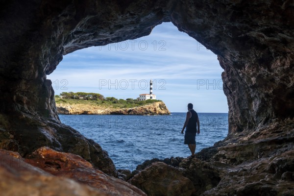 Tourist stands inside the arc natural cave in portocolom, mallorca, admiring the scenic view of a lighthouse across the blue waters