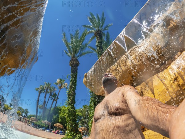 Man refreshing under a waterfall in a water park during summer vacation, enjoying the cool water and the sunny day