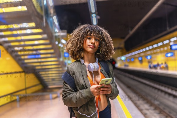 Latin young woman waiting in the platform of underground metro using phone and carrying headphones