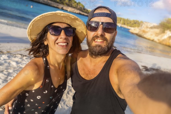 A man and woman are smiling for the camera on a beach. The man is wearing a black tank top and sunglasses, while the woman is wearing a straw hat