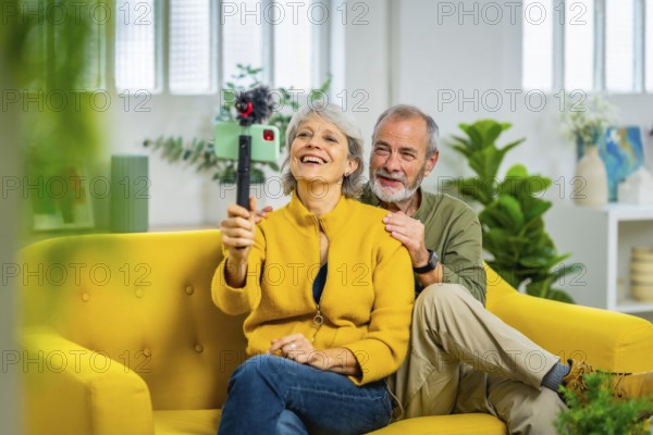 Happy senior couple holding mobile phone during video call sitting on modern yellow sofa at home