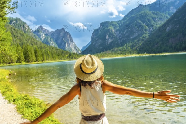 Woman wearing straw hat with open arms enjoying the pristine waters of lake dobbiaco and the majestic dolomites mountains in summer