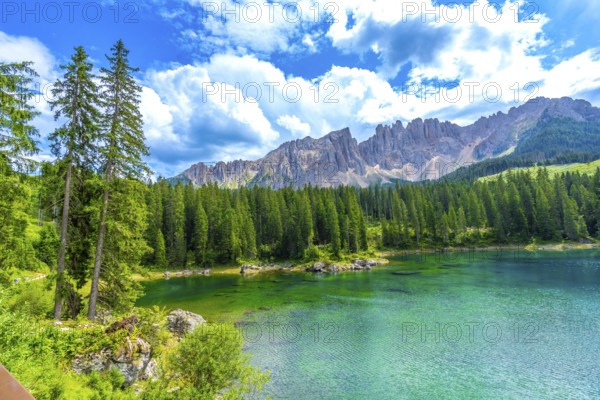 Emerald waters of lake carezza reflecting latemar mountain range, surrounded by lush pine forest under blue sky with white clouds in summer, dolomites, italy