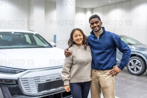 Happy multiethnic couple hugging, buying new electric car at dealership, embracing modern eco friendly transportation