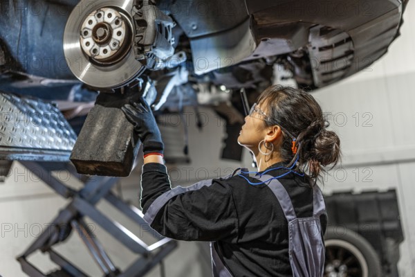 Skilled female mechanic working under a lifted car, inspecting and repairing the brake system in a professional workshop