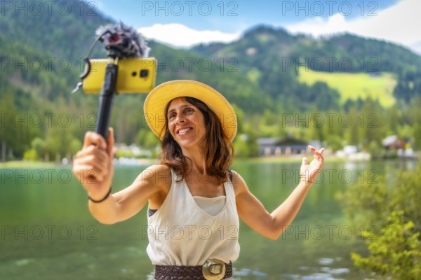 Smiling woman wearing straw hat vlogging with smartphone and external microphone at lake dobbiaco in the italian dolomites during summer holidays