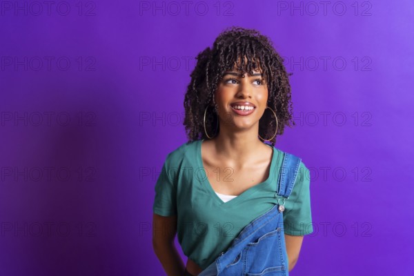 Young woman with curly hair, wearing a green t shirt and denim overalls, smiles while looking up against a vibrant purple backdrop
