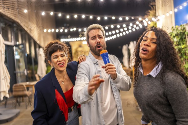 Man and two multiracial women singing karaoke having fun at night party in a terrace of a bar