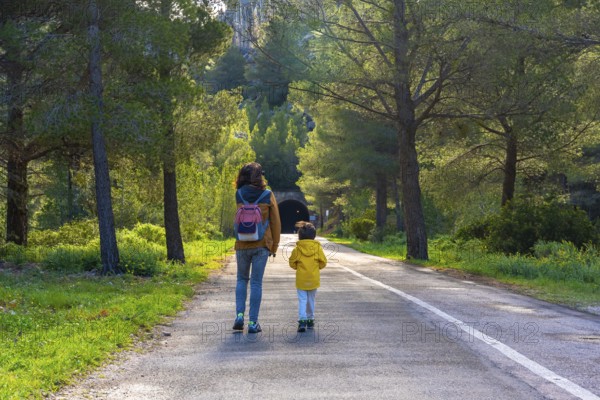 Mother and child enjoying a leisurely walk on a paved road amidst lush greenery and pine trees near a tunnel in la fontcalda sanctuary, gandesa, tarragona