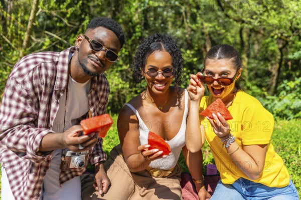 Three joyful friends enjoying fresh watermelon slices while relaxing at a sunny summer picnic in a vibrant park, surrounded by nature