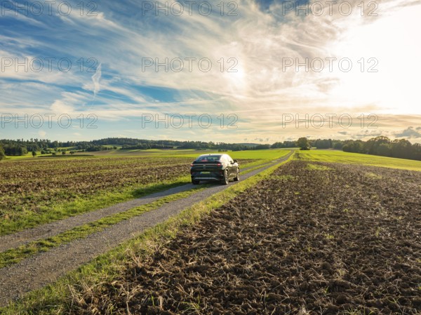 A car drives on a rural road between ploughed fields under a partly cloudy sky, Carsharing, VW ID5, Calw, Black Forest, Germany, Europe
