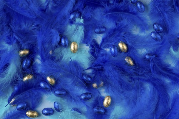 Golden and blue chocolate eggs amidst dense blue feathers create a festive texture