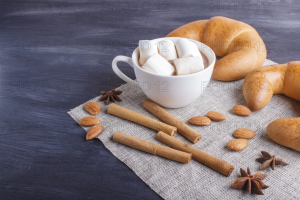A cup of hot chocolate with marshmallow, croissants and spices on linen napkin and black wooden background. close up, copy space
