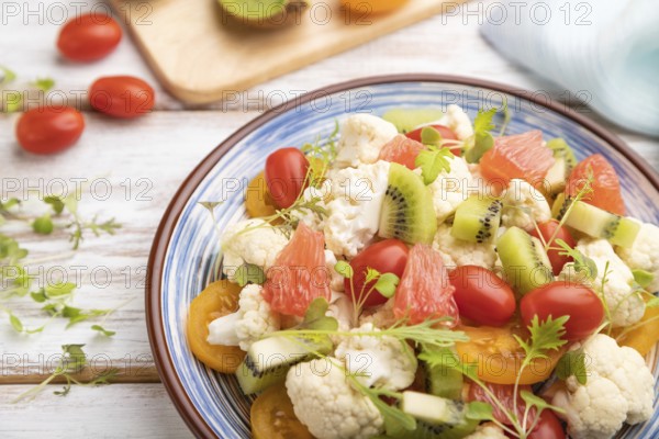 Vegetarian salad of cauliflower cabbage, kiwi, tomatoes, microgreen sprouts on white wooden background and blue linen textile. Side view, close up, selective focus