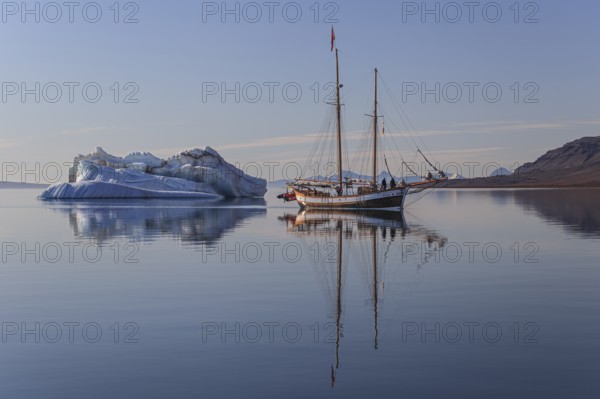 Traditional sailing ship reflected in fjord next to iceberg, sunny, Ittoqqortoormiit, Scoresby Sund, East Greenland, Greenland, North America