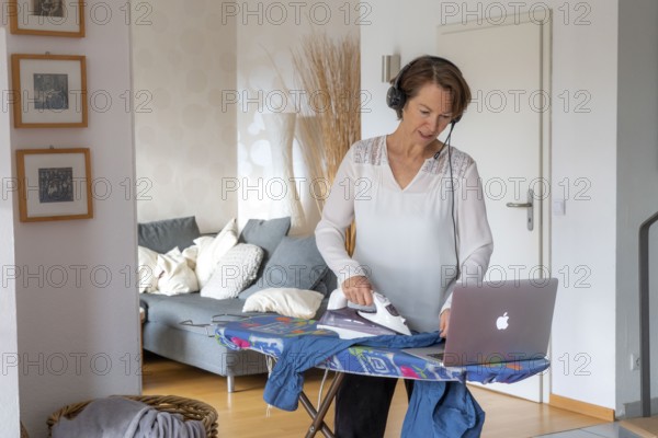 Woman, mid-50s, works from home, with laptop and communicates with colleagues via headset, home office, irons laundry in parallel