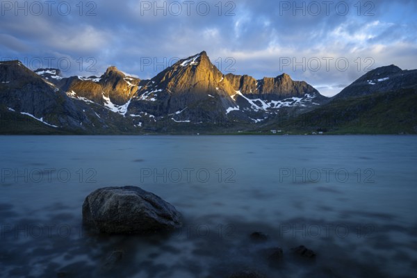 Landscape with sea and mountains on the Lofoten Islands, view over the fjord Flakstadpollen to the mountain Stortinden and others. A rock in the foreground. At night at the time of the midnight sun, some clouds in the sky, the sun shining on the mountain peaks. Long exposure. Early summer. Flakstadoya, Lofoten, Norway, Europe