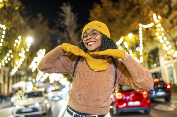 Smiling young woman wearing yellow winter clothes enjoying christmas lights in a city street at night