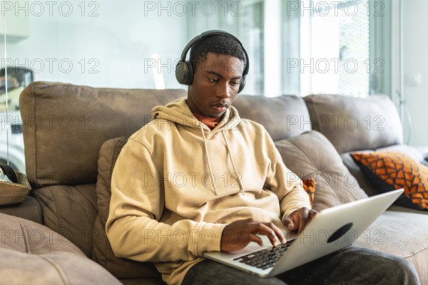 Young black man using laptop and headphones while working or studying from home, sitting on a comfortable couch