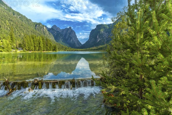 Crystal clear water cascading from a small waterfall into lake dobbiaco, reflecting the majestic dolomite peaks under a summer sky