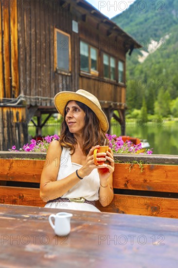 Woman wearing straw hat holding a mug and enjoying a hot beverage at a wooden table with lake dobbiaco and the dolomites in the background