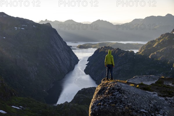 Mountaineer, young man standing on rocks and looking into the distance, at sunrise, fjord Trollfjord and mountains, hike to Trollfjord Hytta, at Trollfjord and Raftsund, Lofoten, Nordland, Norway, Europe