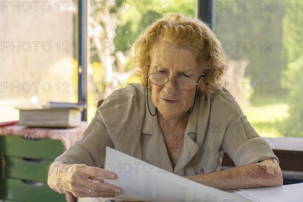Focused elderly woman wearing glasses reading important documents while sitting at her home office desk, concentrating on paperwork and administrative tasks