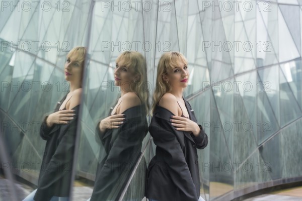 Fashionable pose of a young blonde in a glass buildings in the city, young man in a black suit looking at the camera reflected in the windows