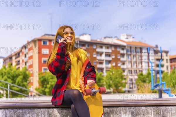 Skater woman in a yellow t-shirt, red plaid shirt and sunglasses, sitting with skateboard in the city, smiling calling with the phone