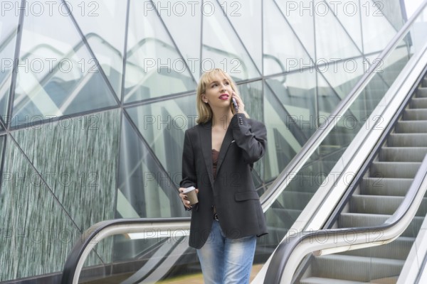 Entrepreneurial girl in business park in a modern building with escalator, young blonde in a suit making a call with a take away coffee