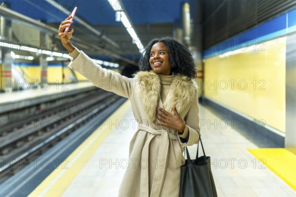 Smiling businesswoman enjoying a selfie with her smartphone while waiting for the subway train at a bustling urban station