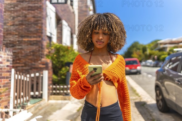 Frontal view of a beauty latin woman with wavy hair and colorful casual clothes using phone walking along a residential district
