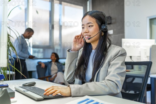 Young asian operator wearing headset working on computer keyboard in a coworking office with colleagues in background