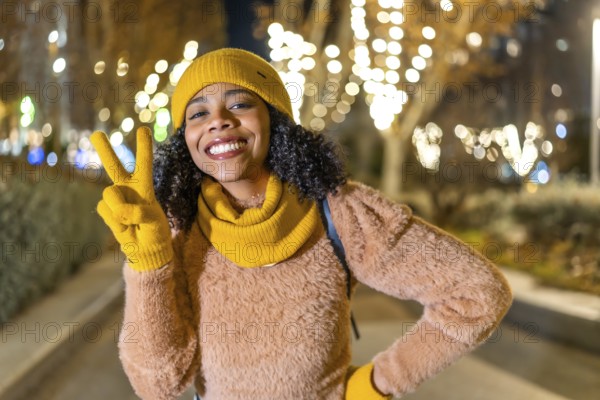 Portrait of a smiling young woman wearing yellow winter clothes showing victory sign with fingers while enjoying christmas illumination in city street at night