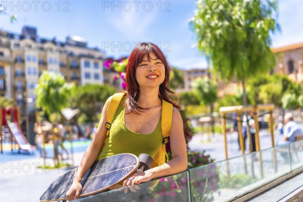 Chinese cool young woman leaning on rail holding skateboard and looking at city from an urban square